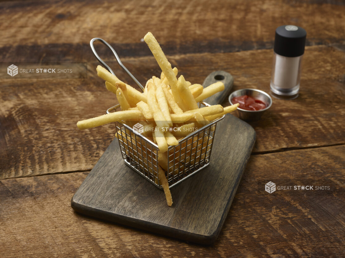 French fries in a small fry basket on a wooden board with a ramekin of ketchup and a salt shaker on a rustic wooden background