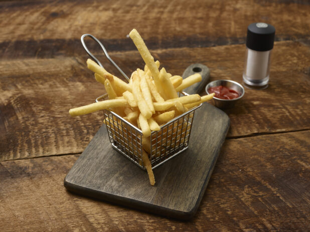 French fries in a small fry basket on a wooden board with a ramekin of ketchup and a salt shaker on a rustic wooden background