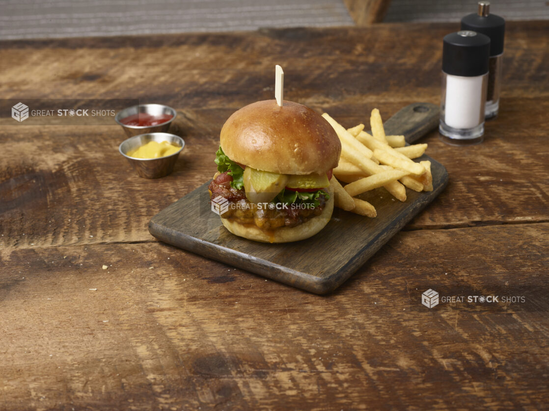 Cheeseburger with pickles, lettuce and tomato and french fries on a wooden board with ramekins of ketchup and mustard with a salt shaker and pepper grinder in the background on a rustic wooden background