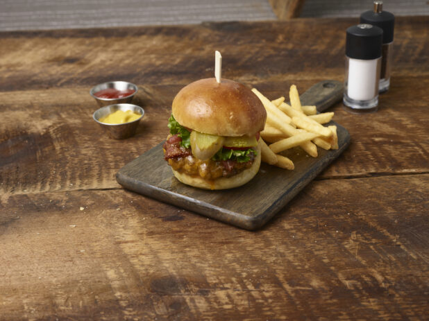 Cheeseburger with pickles, lettuce and tomato and french fries on a wooden board with ramekins of ketchup and mustard with a salt shaker and pepper grinder in the background on a rustic wooden background