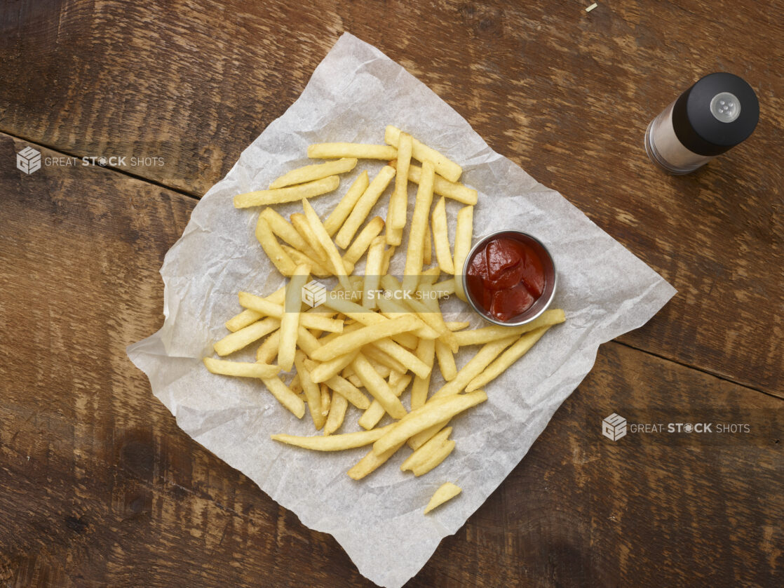 Overhead view of french fries with a ramekin of ketchup on parchment paper with a salt shaker on a rustic wooden background