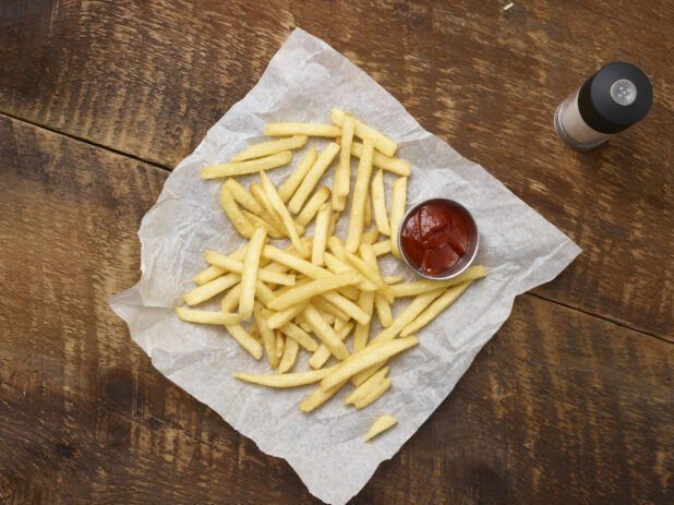 Overhead view of french fries with a ramekin of ketchup on parchment paper with a salt shaker on a rustic wooden background