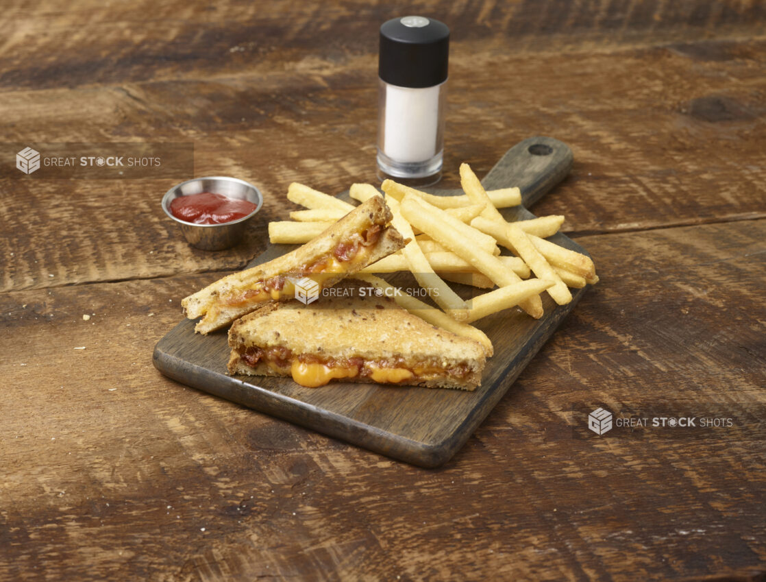 Grilled cheese and bacon sandwich with french fries on a wooden board with a ramekin of ketchup and a salt shaker in the background on a rustic wooden background