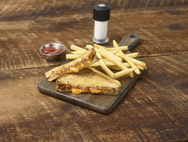 Grilled cheese and bacon sandwich with french fries on a wooden board with a ramekin of ketchup and a salt shaker in the background on a rustic wooden background