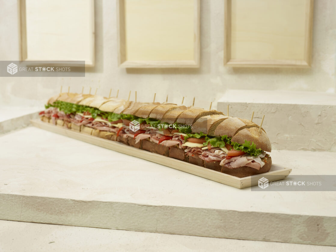 Jumbo-Sized Party Sub Sandwich with Lettuce, Tomato, Cheese and Deli Meats, Sliced on a Long and Narrow Wood Serving Tray on a Beige Surface and Background