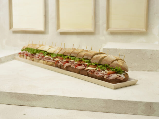 Jumbo-Sized Party Sub Sandwich with Lettuce, Tomato, Cheese and Deli Meats, Sliced on a Long and Narrow Wood Serving Tray on a Beige Surface and Background