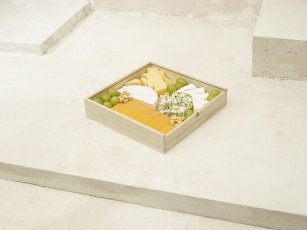 An Assortment of Sliced Cheeses with Fresh Green Grapes in a Square Wood Serving Tray for Catering on a Beige Painted Background