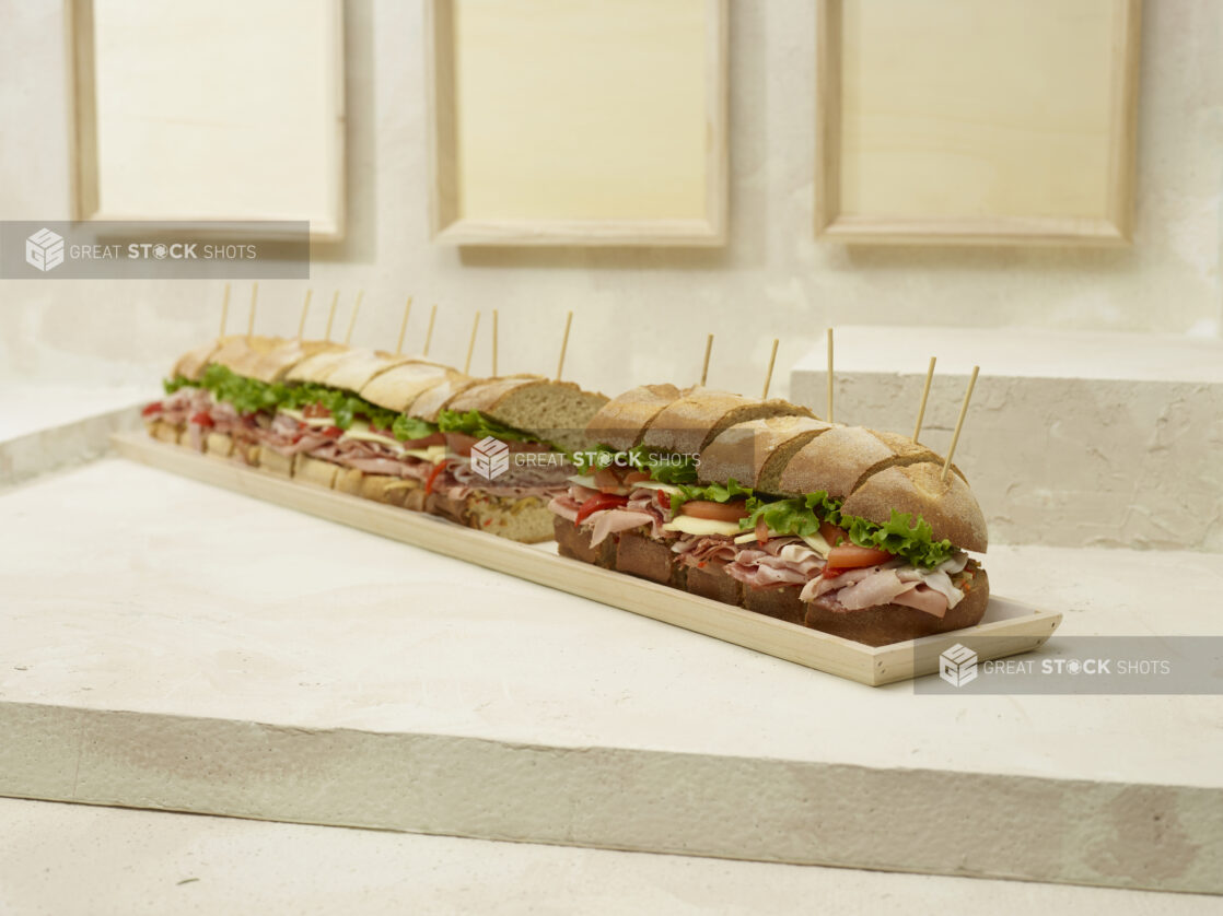 Jumbo-Sized Party Sub Sandwich with Lettuce, Tomato, Cheese and Deli Meats, Sliced on a Long and Narrow Wood Serving Tray on a Beige Surface and Background - Variation