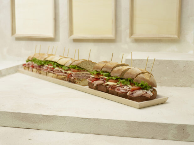Jumbo-Sized Party Sub Sandwich with Lettuce, Tomato, Cheese and Deli Meats, Sliced on a Long and Narrow Wood Serving Tray on a Beige Surface and Background - Variation
