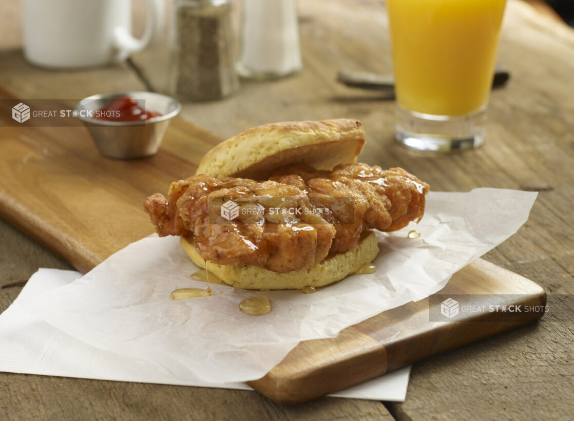 Crispy fried chicken breakfast sandwich with honey on a biscuit on a wooden board with orange juice and ketchup in the background on a rustic wooden background