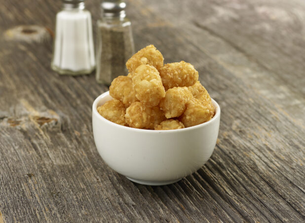 Hash brown potatoes, tater tots, in a small white ceramic bowl with salt and pepper shaker in the background on a rustic wooden background