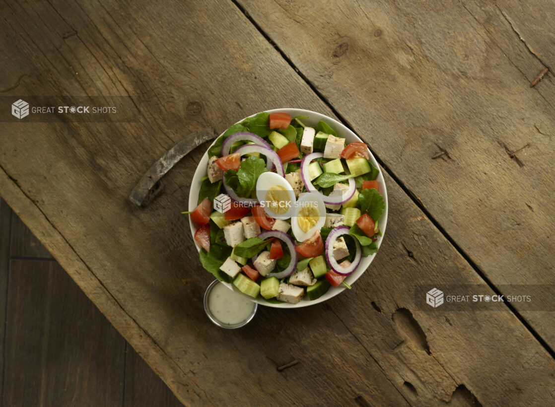 Mixed greens salad with chicken cubes, red bell pepper, red onion and boiled eggs with dressing on the side on a rustic wooden background, overhead view