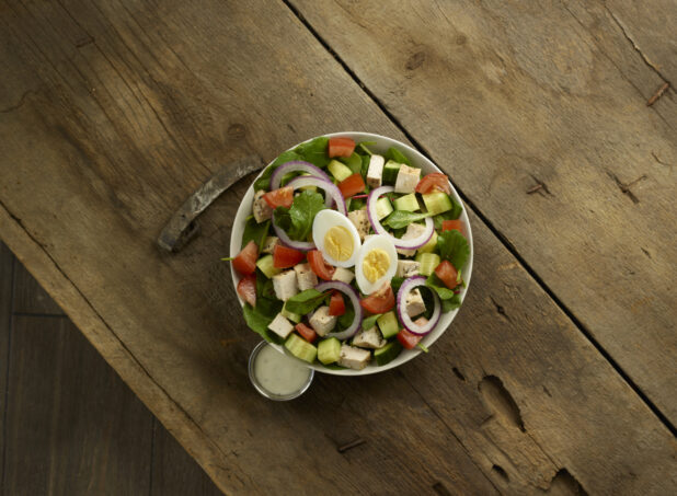 Mixed greens salad with chicken cubes, red bell pepper, red onion and boiled eggs with dressing on the side on a rustic wooden background, overhead view