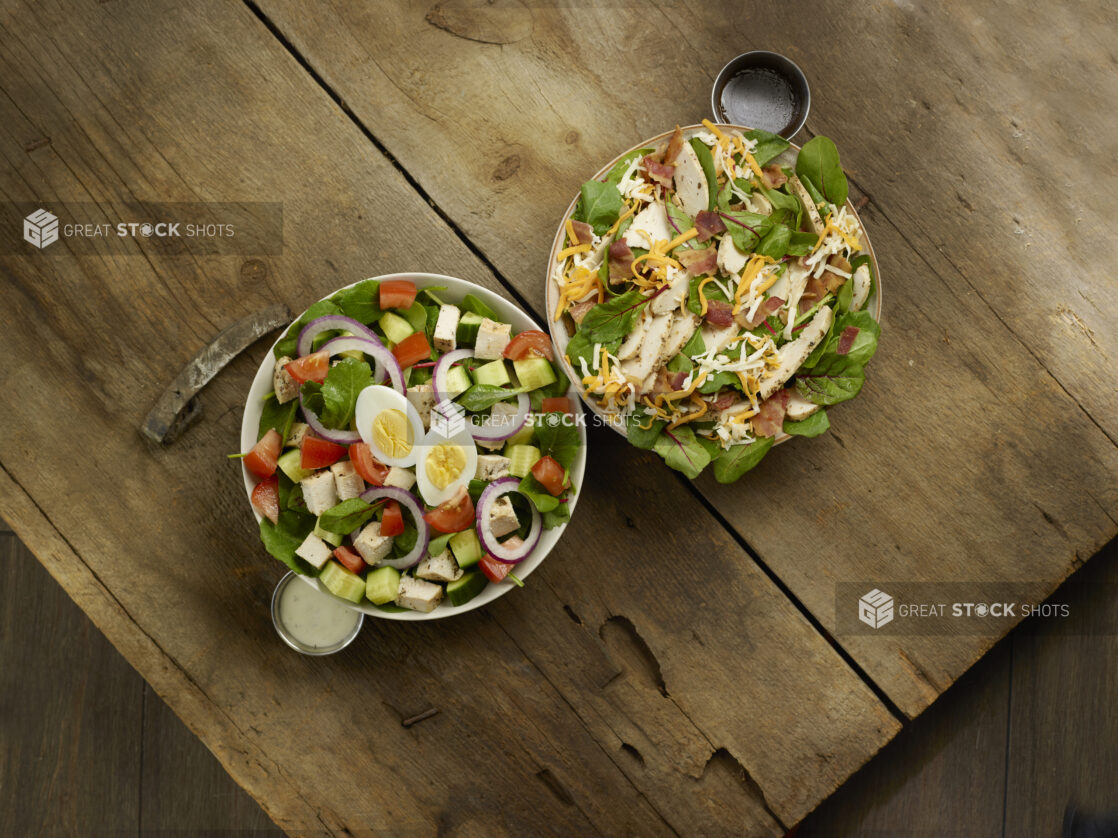 2 salads, mixed greens with chicken cubes, red bell pepper, red onion and boiled eggs, and mixed greens with sliced chicken, bacon and shredded cheddar, dressing on the side all on rustic wooden boards, overhead view