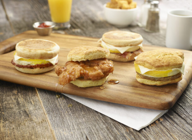 Assorted breakfast sandwiches on a wooden board, with accompaniments in the background on a rustic wooden background