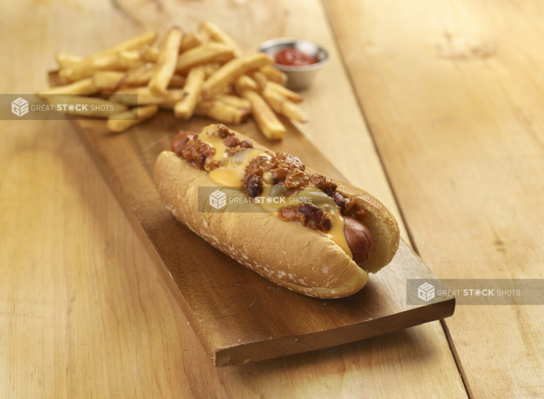 Chili dog with french fries on a wooden board on a wooden table with a small ramekin of ketchup