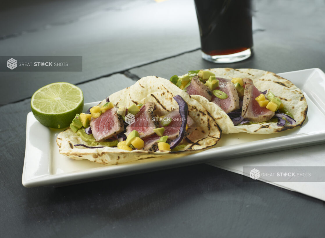 Seared tuna tacos with corn, avocado and red cabbage on a rectangular white plate with a sliced lime and a beverage in the background