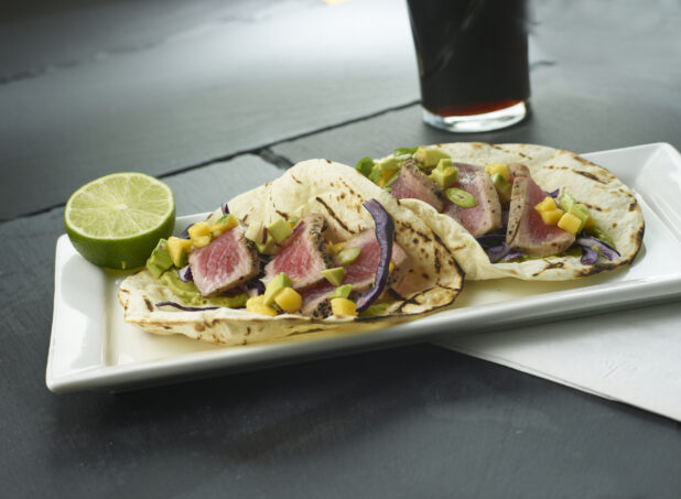 Seared tuna tacos with corn, avocado and red cabbage on a rectangular white plate with a sliced lime and a beverage in the background
