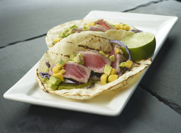 2 seared tuna tacos in grilled soft taco shells with fresh mango, avocado, red cabbage and lime on a white rectangular plate, close up view