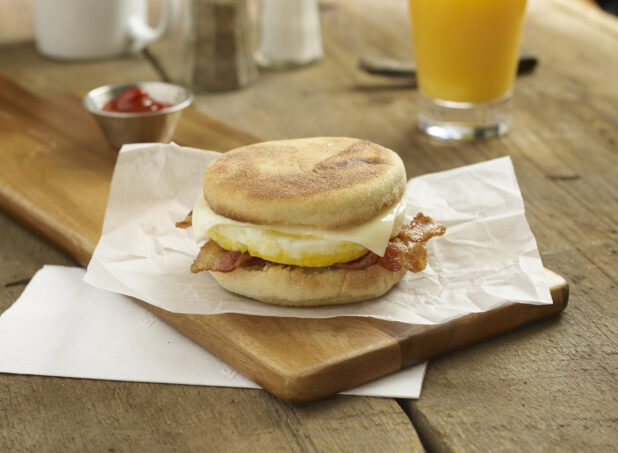 Egg and bacon breakfast sandwich cheese on an english muffin on a wood board with a glass of orange juice and ketchup in the background on a rustic wooden background