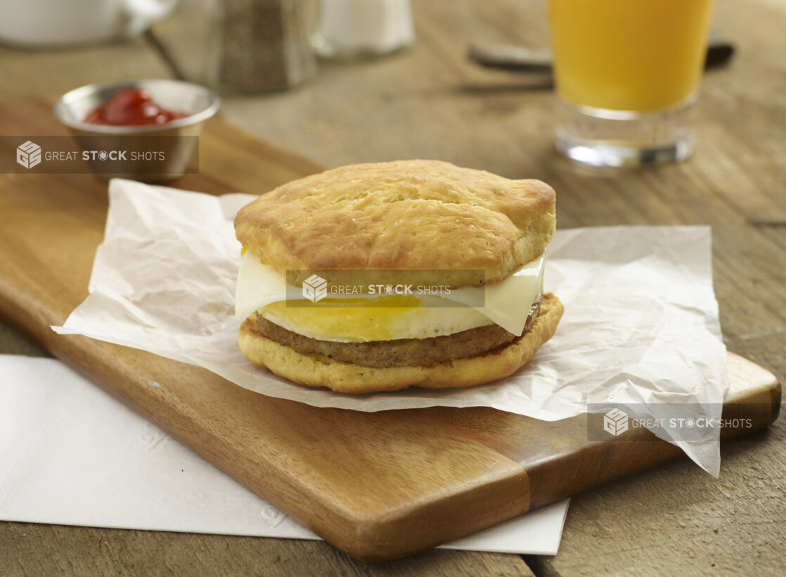 Egg and sausage breakfast sandwich with cheese on a biscuit on a wooden board with orange juice and ketchup in a ramekin in the background on a rustic wooden background