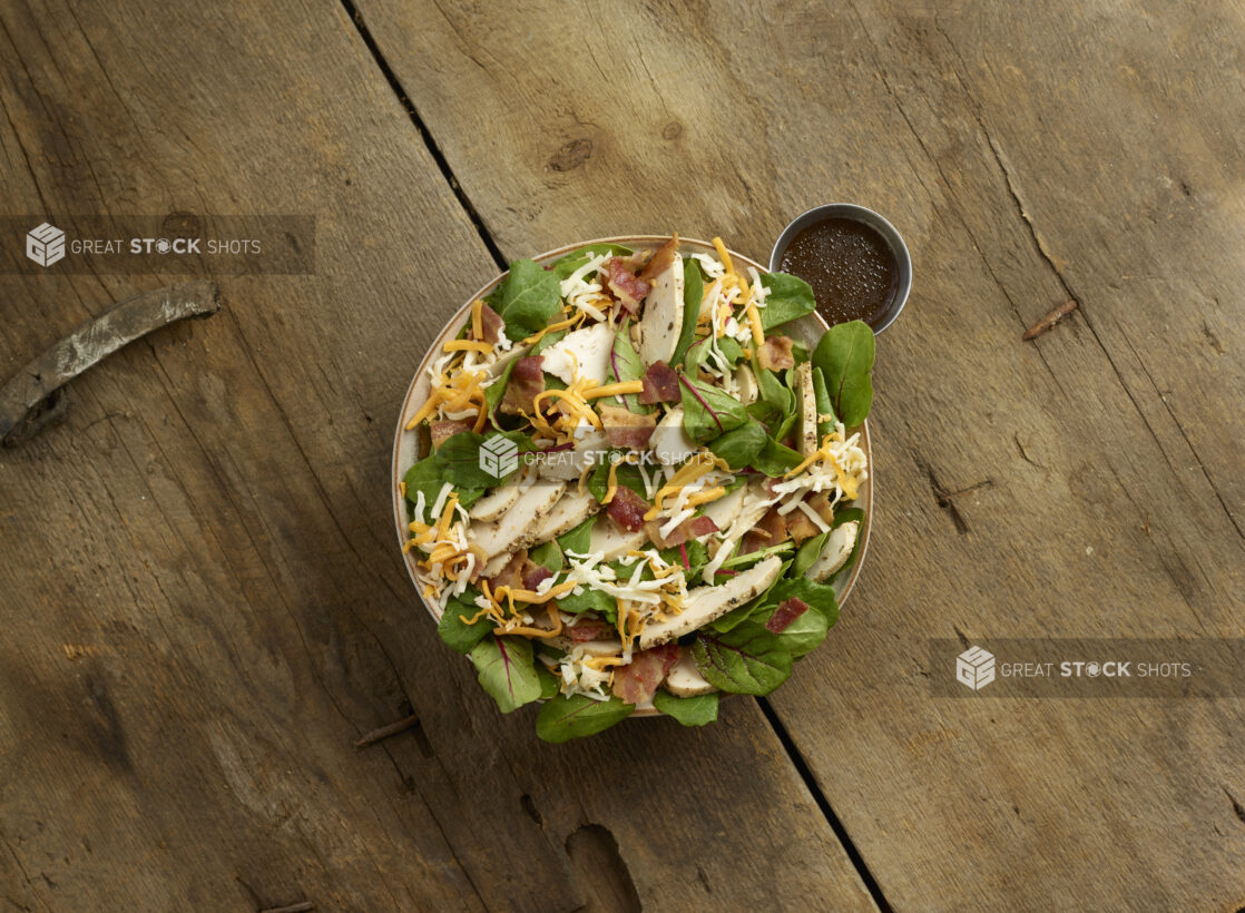 Overhead view of a mixed greens salad with sliced chicken, bacon and shredded cheddar cheese in a bowl with dressing on the side on a rustic wooden background