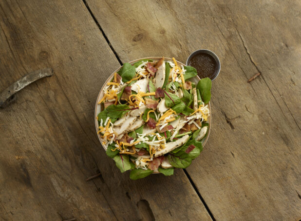 Overhead view of a mixed greens salad with sliced chicken, bacon and shredded cheddar cheese in a bowl with dressing on the side on a rustic wooden background
