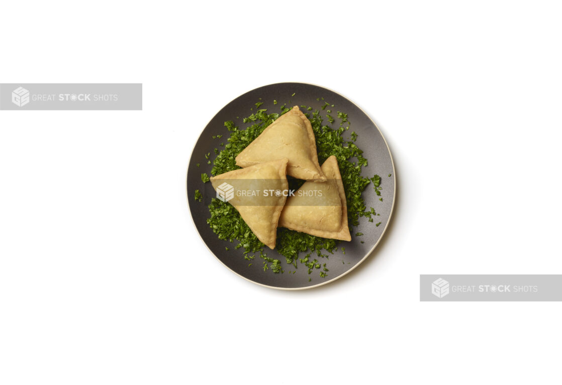 Overhead View of Vegetarian Samosas on a Bed of Chopped Fresh Parsley and Round Black Ceramic Dish, on a White Background for Isolation