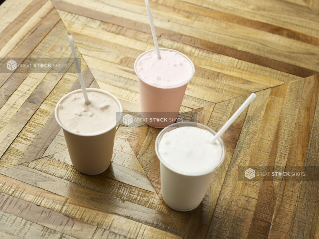 Strawberry, Chocolate and Vanilla Milkshakes in Clear Plastic Cups with Plastic Straws on a Parquet Wooden Floor in an Indoor Setting