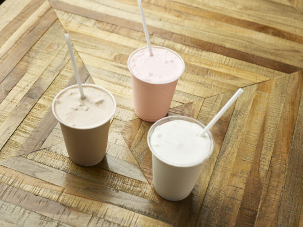 Strawberry, Chocolate and Vanilla Milkshakes in Clear Plastic Cups with Plastic Straws on a Parquet Wooden Floor in an Indoor Setting