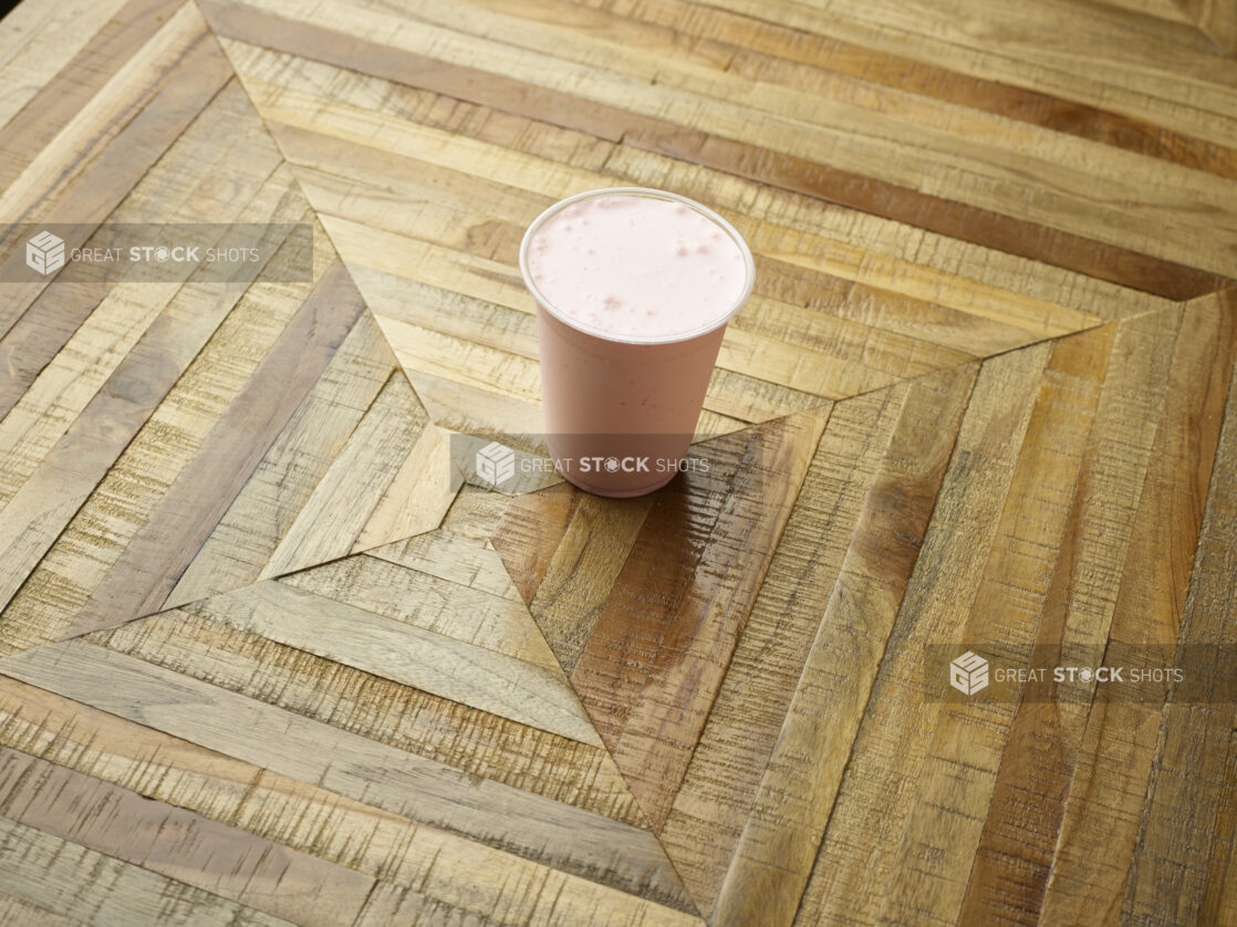 Strawberry Milkshake in a Clear Plastic Cup on a Parquet Wooden Tabletop in an Indoor Setting