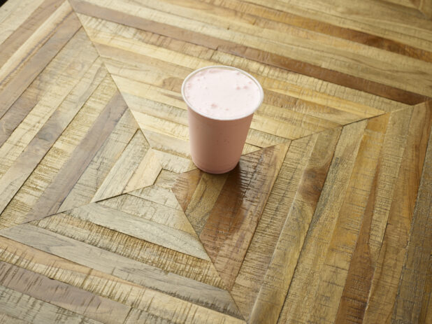 Strawberry Milkshake in a Clear Plastic Cup on a Parquet Wooden Tabletop in an Indoor Setting