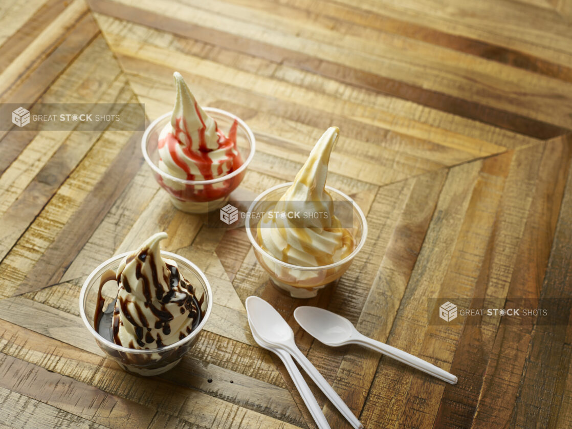 Strawberry, Chocolate and Caramel Ice Cream Sundaes in Clear Plastic Cups with White Plastic Spoons on a Parquet Wooden Floor in an Indoor Setting