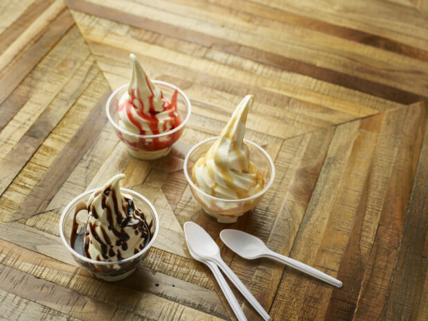 Strawberry, Chocolate and Caramel Ice Cream Sundaes in Clear Plastic Cups with White Plastic Spoons on a Parquet Wooden Floor in an Indoor Setting
