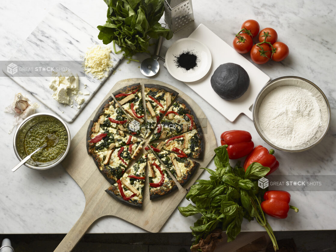 Overhead View of a Sliced Specialty Charcoal Crust Pizza with Grilled Chicken, Spinach and Red Peppers on a Wooden Pizza Peel, Surrounded by Fresh Ingredients on a Marble Counter Top