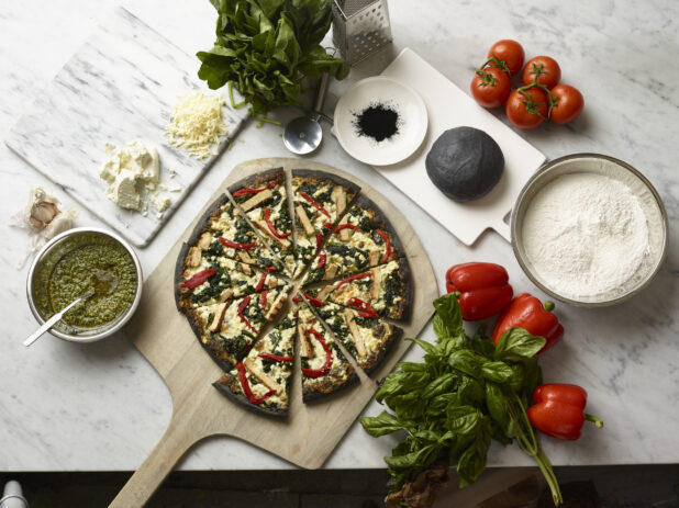 Overhead View of a Sliced Specialty Charcoal Crust Pizza with Grilled Chicken, Spinach and Red Peppers on a Wooden Pizza Peel, Surrounded by Fresh Ingredients on a Marble Counter Top