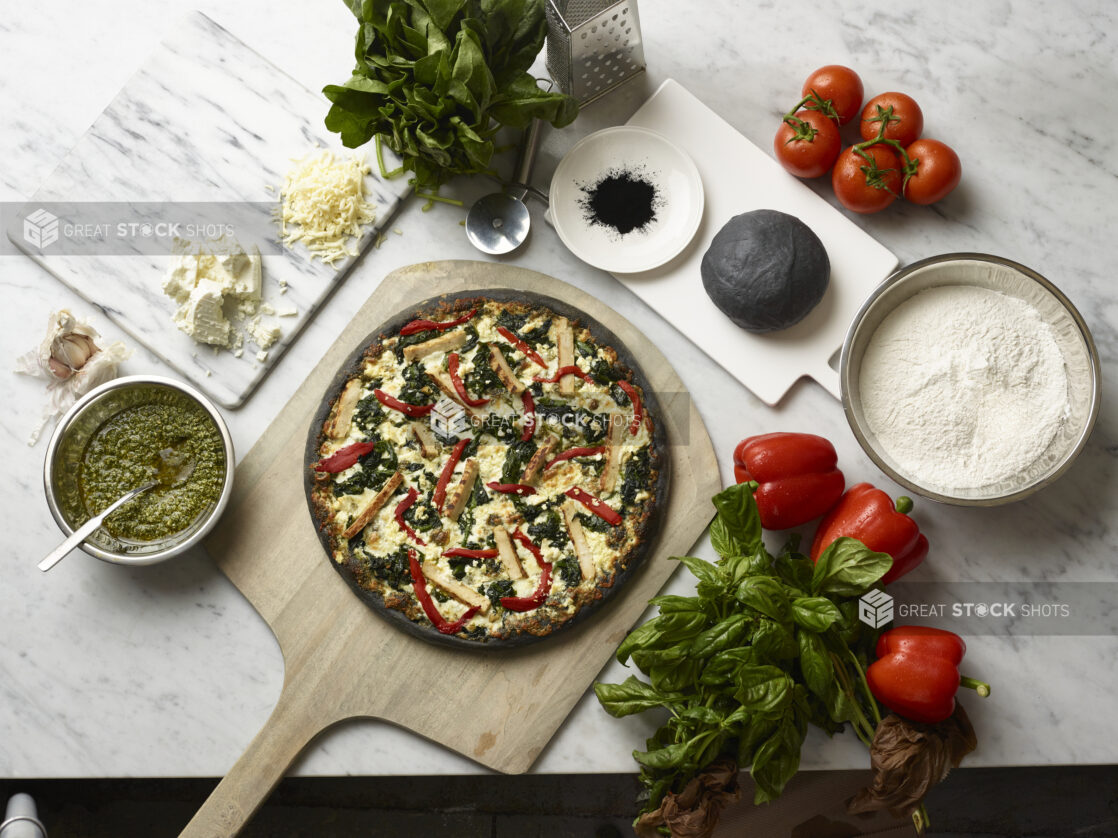 Overhead View of a Specialty Charcoal Crust Pizza with Grilled Chicken, Spinach and Red Peppers on a Wooden Pizza Peel, Surrounded by Fresh Ingredients on a Marble Counter Top