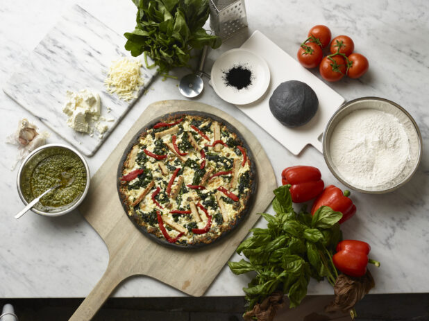 Overhead View of a Specialty Charcoal Crust Pizza with Grilled Chicken, Spinach and Red Peppers on a Wooden Pizza Peel, Surrounded by Fresh Ingredients on a Marble Counter Top