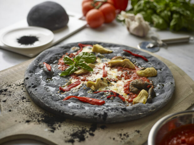 Close Up of a Personal Pizza with Charcoal Infused Pizza Crust, Roasted Red Peppers, Artichoke Hearts and Fresh Basil Leaves on a Wooden Pizza Peel with Fresh Ingredients in the Background