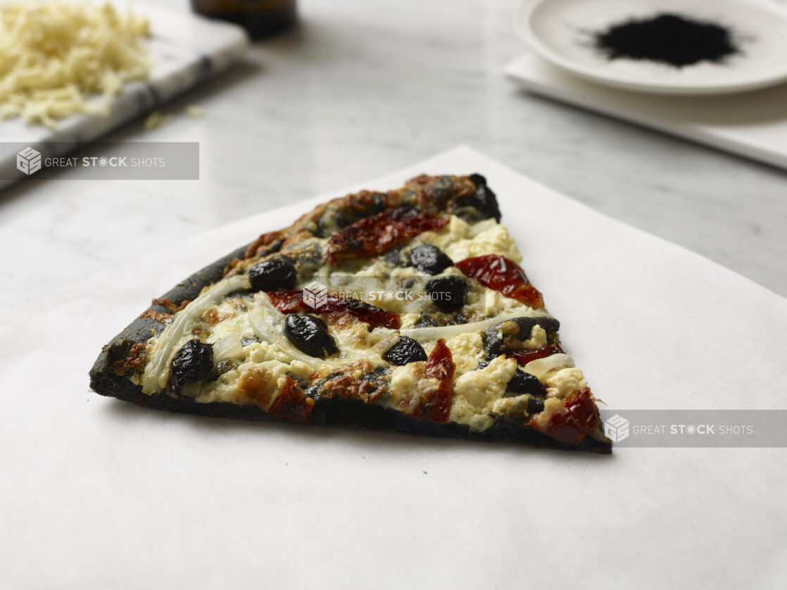 Slice of Pizza with a Charcoal Infused Pizza Crust, Black Olives, White Onions and Sun-dried Tomato Toppings on a Marble Counter Surface