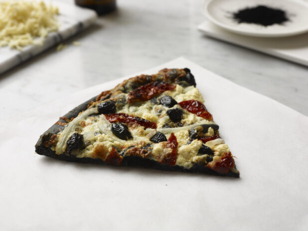 Slice of Pizza with a Charcoal Infused Pizza Crust, Black Olives, White Onions and Sun-dried Tomato Toppings on a Marble Counter Surface