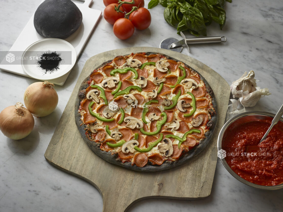 Overhead View of a Specialty Charcoal Crust Deluxe Pizza with Pepperoni, Green Peppers and Mushrooms Toppings on a Wooden Pizza Peel, Surrounded by Fresh Ingredients on a Marble Counter Top