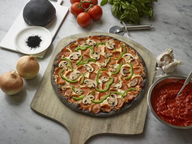 Overhead View of a Specialty Charcoal Crust Deluxe Pizza with Pepperoni, Green Peppers and Mushrooms Toppings on a Wooden Pizza Peel, Surrounded by Fresh Ingredients on a Marble Counter Top