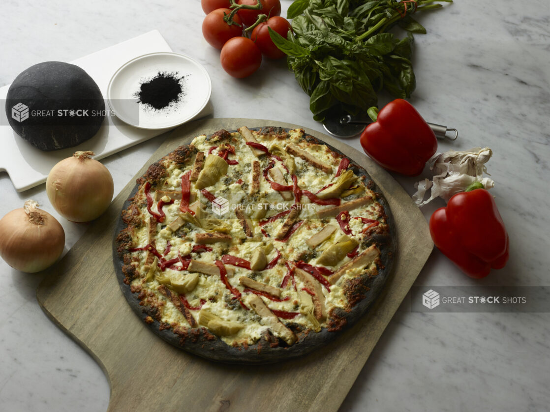 Overhead View of a Specialty Charcoal Crust Pizza with Grilled Chicken, Roasted Red Peppers, Feta Cheese and Artichoke Hearts Toppings on a Wooden Pizza Peel, Surrounded by Fresh Ingredients on a Marble Counter Top