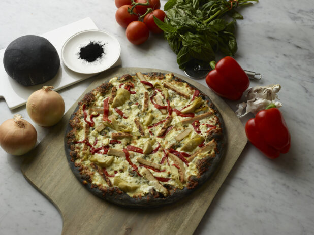 Overhead View of a Specialty Charcoal Crust Pizza with Grilled Chicken, Roasted Red Peppers, Feta Cheese and Artichoke Hearts Toppings on a Wooden Pizza Peel, Surrounded by Fresh Ingredients on a Marble Counter Top