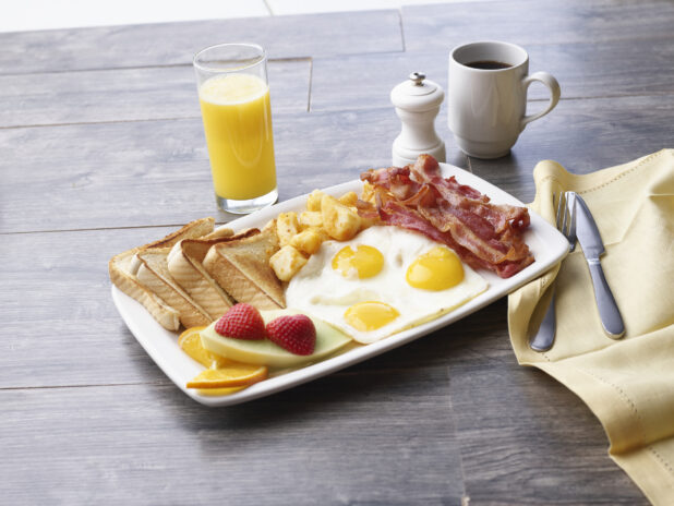 3 eggs sunny side up, breakfast sausages, homefries, white toast, and fruit on a white platter alongside coffee and juice on a gray wooden background