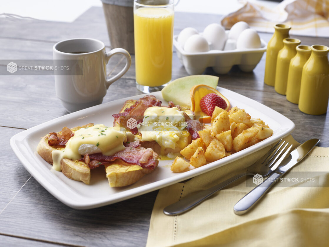 A breakfast plate of Bacon Eggs Benedict on toast with homefries and fruit garnish, breakfast cooking items in background