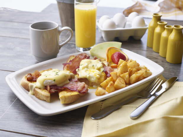 A breakfast plate of Bacon Eggs Benedict on toast with homefries and fruit garnish, breakfast cooking items in background