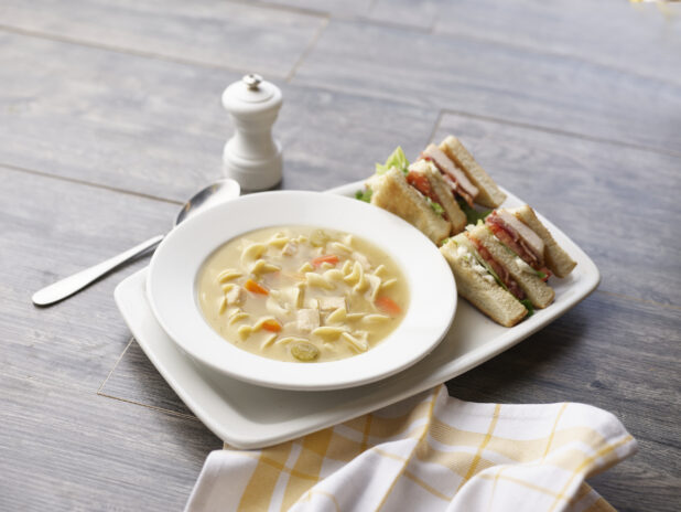 Chicken noodle soup with a half clubhouse sandwich on a grey wood tabletop