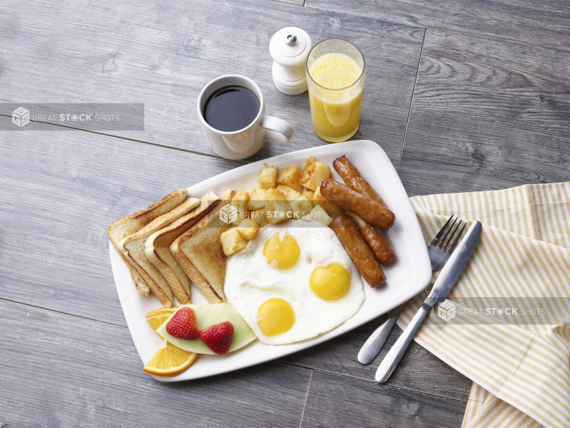 3 eggs sunny side up, breakfast sausages, breakfast potatoes, white toast, and fruit on a white platter alongside coffee and juice on a gray wooden background